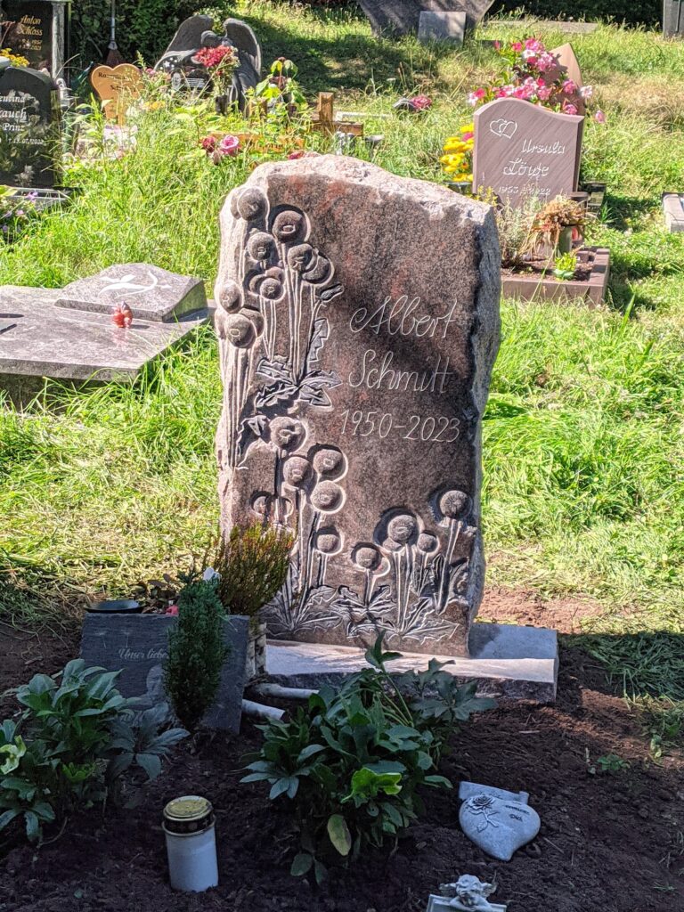 Ein Grabstein, der aus einem alten Grabstein aus Granit gearbeitet wurde. Es zeigt eine Pusteblumen-Wiese, die als Ornament angelegt ist.
